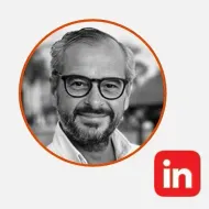 Una foto en escala de grises de un hombre sonriente con gafas en un marco circular, con el logotipo de LinkedIn en la parte inferior derecha.